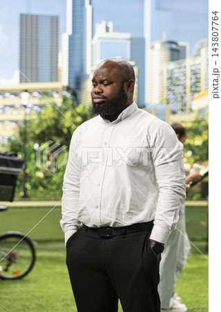 Disappointed businessman standing with hands in pockets and looking downward in urban park, expressing quiet moment of concern against skyline. Professional setback. Disappointed businessman standing with hands in pockets and looking downward in urban park, expressing quiet moment of concern against skyline. Professional setback. 143807764