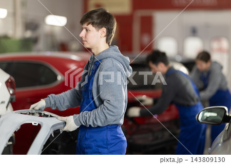 Young auto mechanic carries freshly painted bumper in his hands to be installed on car Young auto mechanic carries freshly painted bumper in his hands to be installed on car 143809130
