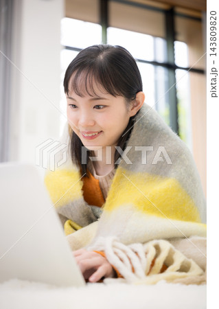 冬の自宅で暖かいストールやブランケットにくるまりながらパソコンを使う笑顔の若い日本人女性 143809820