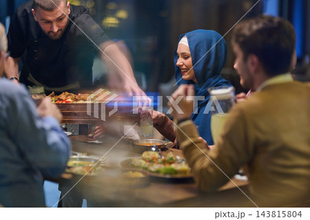 Smiling Woman in Hijab Enjoying Group Dinner at Restaurant While Server Presents Shared Platter 143815804