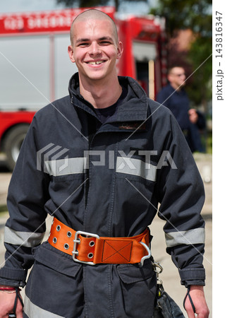Smiling Firefighter in Protective Gear With Bright Safety Belt Standing by Fire Truck 143816347