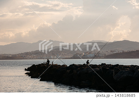 Silhouettes of fishermen on a rocky breakwater in Ceuta with the coast of Morocco at sunset Silhouettes of fishermen on a rocky breakwater in Ceuta with the coast of Morocco at sunset 143818685