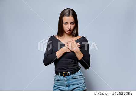 Serious young woman holding hands on her chest on gray background 143820098