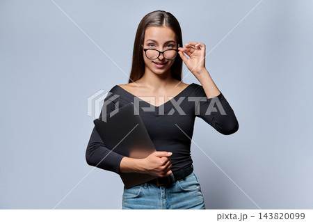 Young woman holding closed laptop and adjusting glasses in studio shot Young woman holding closed laptop and adjusting glasses in studio shot 143820099