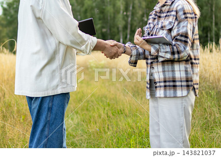 Faceless agricultural partners shake hands in a golden wheat field while holding digital tablets. Concept of business agreement, successful deal and agricultural collaboration 143821037