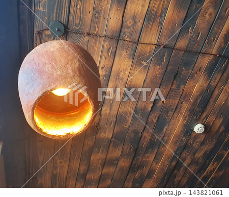 A pendant light with a warm glow and a textured terracotta shade, mounted beneath a ceiling made of wooden planks. A pendant light with a warm glow and a textured terracotta shade, mounted beneath a ceiling made of wooden planks. 143821061