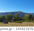 Autumn mountain landscape with forested hills under a clear blue sky, old wooden barn on a dry grass meadow and cozy log houses among birch and spruce trees 143821076