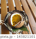 Tropical dessert served in a fresh coconut shell with passion fruit puree and seeds, set on a coconut bowl with straw and a green leaf on a wooden bench, top view 143821101