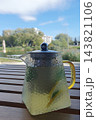 Glass teapot with refreshing lemon and mint herbal tea on a wooden table at an outdoor cafe, blurred lake and park with trees in the background 143821106