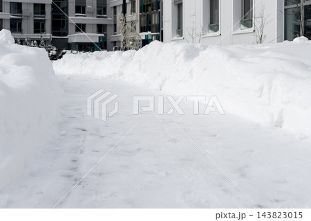 Snowdrift banks line snow covered path, shoveled walkway cleared for winter walk. Footprints and trail on snowy pile near apartment buildings. Urban residential district, overcast cityscape. Snowdrift banks line snow covered path, shoveled walkway cleared for winter walk. Footprints and trail on snowy pile near apartment buildings. Urban residential district, overcast cityscape. 143823015
