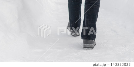 Legs and boots walking through snow, person on snowy winter path. Black jeans and footwear on snow covered walkway, cropped rear view near snowbank. Footsteps on trail, cold wintertime Legs and boots walking through snow, person on snowy winter path. Black jeans and footwear on snow covered walkway, cropped rear view near snowbank. Footsteps on trail, cold wintertime 143823025