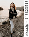 Woman Coastline Fashion Stylish woman in a black leather jacket and striped sweater posing on a pebble beach at sunset 143823513