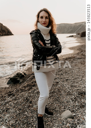 Woman Coastline Fashion Stylish woman in a black leather jacket and striped sweater posing on a pebble beach at sunset Woman Coastline Fashion Stylish woman in a black leather jacket and striped sweater posing on a pebble beach at sunset 143823513