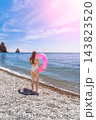 Woman beach swimsuit standing on pebble shore with pink inflatable ring enjoying summer vacation by blue ocean waters under bright sun 143823520