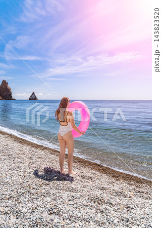 Woman beach swimsuit standing on pebble shore with pink inflatable ring enjoying summer vacation by blue ocean waters under bright sun Woman beach swimsuit standing on pebble shore with pink inflatable ring enjoying summer vacation by blue ocean waters under bright sun 143823520