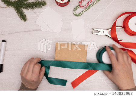 Hands wrap a kraft gift box with green ribbon at a festive Christmas gift station Hands wrap a kraft gift box with green ribbon at a festive Christmas gift station 143823968