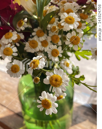 A beautiful bouquet of small white chamomile flowers with golden yellow centers arranged in a translucent green glass vase on a wooden table. Close-up floral photography, rustic home decor A beautiful bouquet of small white chamomile flowers with golden yellow centers arranged in a translucent green glass vase on a wooden table. Close-up floral photography, rustic home decor 143824708