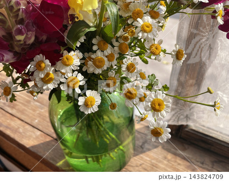 A beautiful bouquet of small white chamomile flowers with golden yellow centers arranged in a translucent green glass vase on a wooden table. Close-up floral photography, rustic home decor A beautiful bouquet of small white chamomile flowers with golden yellow centers arranged in a translucent green glass vase on a wooden table. Close-up floral photography, rustic home decor 143824709