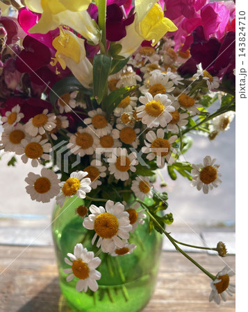 A beautiful bouquet of small white chamomile flowers with golden yellow centers arranged in a translucent green glass vase on a wooden table. Close-up floral photography, rustic home decor  143824710