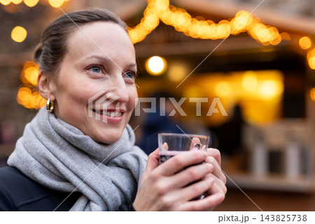 Smiling woman enjoy drink warm mulled wine at Christmas market, wearing black knit hat and gray scarf. Golden festive lights create cozy winter atmosphere during cheerful holiday outing in Germany Smiling woman enjoy drink warm mulled wine at Christmas market, wearing black knit hat and gray scarf. Golden festive lights create cozy winter atmosphere during cheerful holiday outing in Germany 143825738