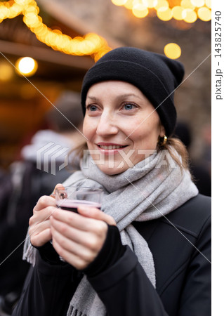 Smiling woman enjoy drink warm mulled wine at Christmas market, wearing black knit hat and gray scarf. Golden festive lights create cozy winter atmosphere during cheerful holiday outing in Germany 143825740