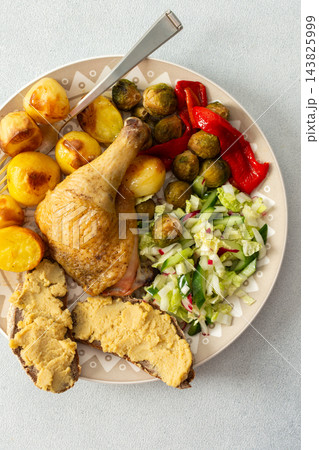 A plate holds baked chicken leg, potatoes, fresh salad, roasted Brussels sprouts, and chickpea bread. This meal is ready to eat at a dining table 143825999