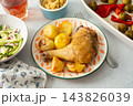 A baked chicken leg rests on a plate with potatoes. Fresh salad, roasted Brussels sprouts, and chickpea bread are arranged on the table. It looks like a wholesome meal 143826039
