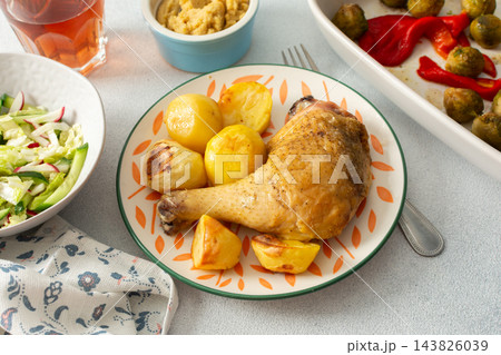 A baked chicken leg rests on a plate with potatoes. Fresh salad, roasted Brussels sprouts, and chickpea bread are arranged on the table. It looks like a wholesome meal A baked chicken leg rests on a plate with potatoes. Fresh salad, roasted Brussels sprouts, and chickpea bread are arranged on the table. It looks like a wholesome meal 143826039