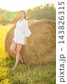 Young woman in white outfit relaxing beside a hay bale in summer field. Rural lifestyle represents slow living, outdoor leisure and connection with nature 143826315