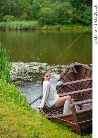 Young woman relaxing in wooden rowboat by lake on autumn day. Cozy sweater and rubber boots represent seasonal outdoor lifestyle and slow living in nature 143826316