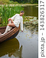 Young woman in white sweater sits on wooden boat, reaching towards water surface surrounded by lily pads and lush greenery in serene lake setting 143826317