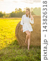 Young woman in white outfit relaxing beside a hay bale in summer field. Rural lifestyle represents slow living, outdoor leisure and connection with nature 143826318