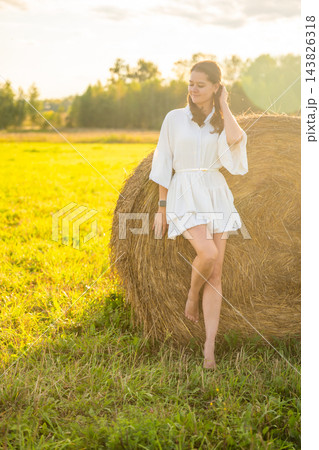 Young woman in white outfit relaxing beside a hay bale in summer field. Rural lifestyle represents slow living, outdoor leisure and connection with nature 143826318