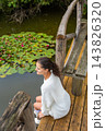 Young woman with dark hair wearing a white sweater sits on wooden dock beside a pond with lily pads and pink flowers in a serene natural setting 143826320