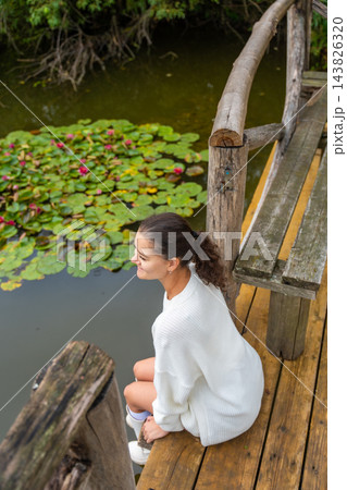 Young woman with dark hair wearing a white sweater sits on wooden dock beside a pond with lily pads and pink flowers in a serene natural setting Young woman with dark hair wearing a white sweater sits on wooden dock beside a pond with lily pads and pink flowers in a serene natural setting 143826320