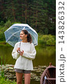 Young woman with curly hair holding a transparent umbrella stands by a serene pond surrounded by lush greenery and a wooden boat in the background 143826332