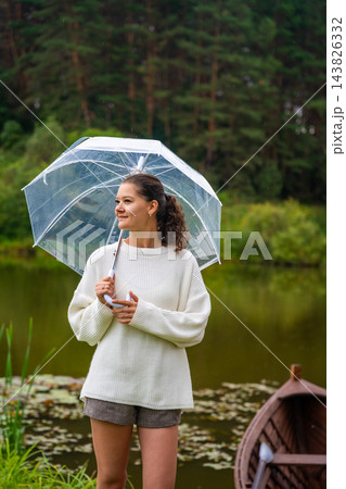 Young woman with curly hair holding a transparent umbrella stands by a serene pond surrounded by lush greenery and a wooden boat in the background Young woman with curly hair holding a transparent umbrella stands by a serene pond surrounded by lush greenery and a wooden boat in the background 143826332