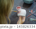 Woman painting pink glaze on handmade ceramic mug with brush in pottery workshop. Pottery decoration represents creativity and learning a craft as a hobby 143826334