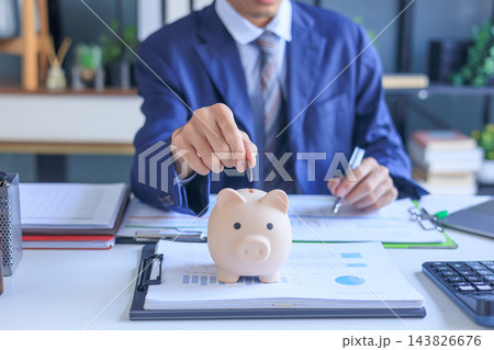 Businessman putting coin into piggy bank on office desk. Concept of saving money. Businessman putting coin into piggy bank on office desk. Concept of saving money. 143826676