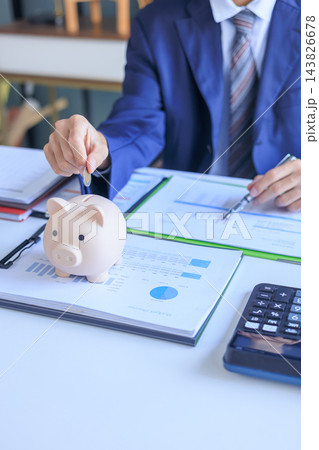 Businessman putting coin into piggy bank on office desk. Concept of saving money. Businessman putting coin into piggy bank on office desk. Concept of saving money. 143826678