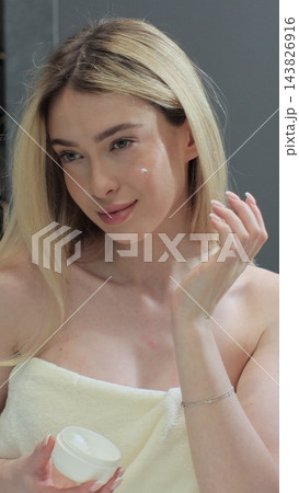 Happy young woman apply skincare cream on face look in bathroom mirror. Facial cream, healthy skin care treatment concept. 143826916