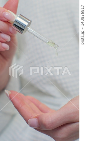 Woman in bathrobe applying hyaluronic collagen vital drops on hands. Skincare routine after morning showering. 143826917