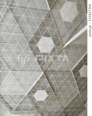 Repeated triangular ceiling members surround pale polygonal panels, creating a delicate overhead pattern within a spacious building Repeated triangular ceiling members surround pale polygonal panels, creating a delicate overhead pattern within a spacious building 143827304
