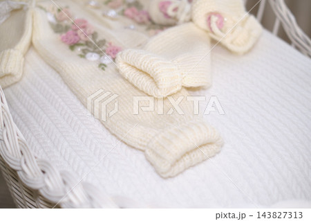A cream knitted baby outfit lies in a white wicker cradle, showing soft cuffs and floral details. The image reflects newborn preparation, handmade care, family tenderness, and gentle anticipation. 143827313