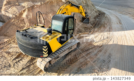 A yellow tracked excavator digs into a cut in the earth beside a dirt road, with deep track marks across the construction site A yellow tracked excavator digs into a cut in the earth beside a dirt road, with deep track marks across the construction site 143827349