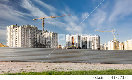 Apartment buildings and cranes stand at a construction site behind a long metal fence, with bare ground in the foreground during daytime 143827350