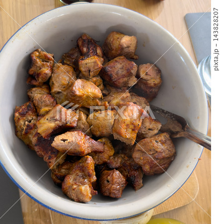 Close-up of freshly grilled pork pieces with a golden-brown crust and charred edges, served in a white enamel bowl with a metal fork. R 143828207