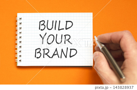 Build your brand words symbol. Concept words Build your brand on beautiful white note. Beautiful orange background. Businessman hand. Business and build your brand concept. Copy space. 143828937
