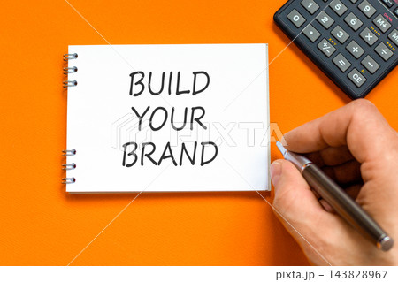 Build your brand words symbol. Concept words Build your brand on beautiful white note. Beautiful orange background. Businessman hand. Business and build your brand concept. Copy space. 143828967
