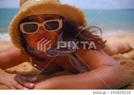 A young woman lies sunbathing on the beach, wearing a straw sun hat and white sunglasses. A young woman lies sunbathing on the beach, wearing a straw sun hat and white sunglasses. 143829216